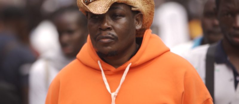 A close-up portrait of a man wearing a bright orange hoodie and a straw hat, looking off-camera with a serious expression. Other people are blurred in the background.